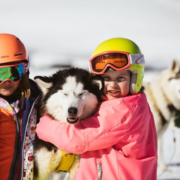 Zwei Kinder mit Skihelmen kuscheln mit einem Husky im Schnee.