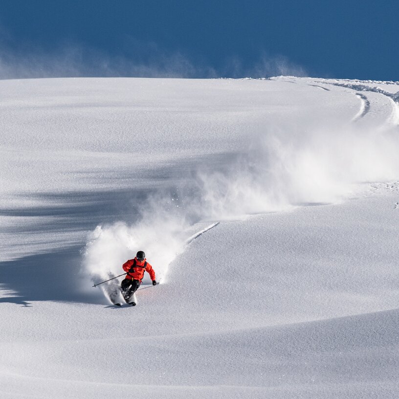 Skifahrer in roter Jacke fährt schwungvoll durch frischen Pulverschnee.