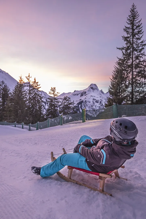 Person sledding on snowy Alpine path at sunset.