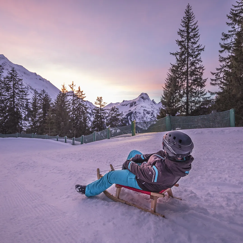 Person fährt mit Schlitten in verschneiter Berglandschaft Richtung Sonnenuntergang.