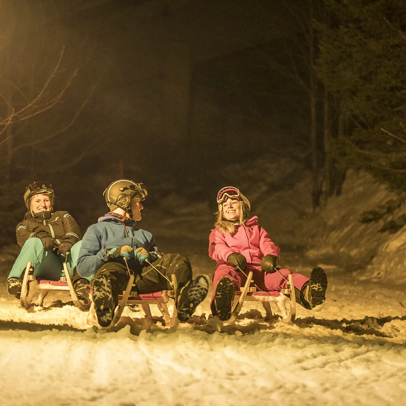 Drei Personen beim nächtlichen Rodeln auf einer verschneiten Rodelpiste.