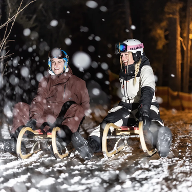 Zwei Personen beim Nachtschlittenfahren durch den verschneiten Wald.