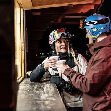 Zwei Personen in Skiausrüstung trinken nachts zusammen an einem Skihüttenfenster ein warmes Getränk.