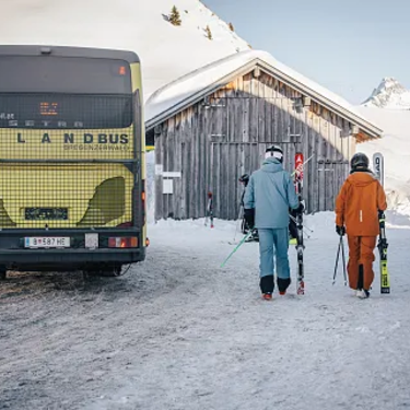 Zwei Skifahrer gehen mit Ausrüstung im Schnee zu einem gelben Skibus.