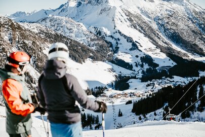Zwei Skifahrer blicken von der Piste aus auf ein verschneites Tal in den Bergen.