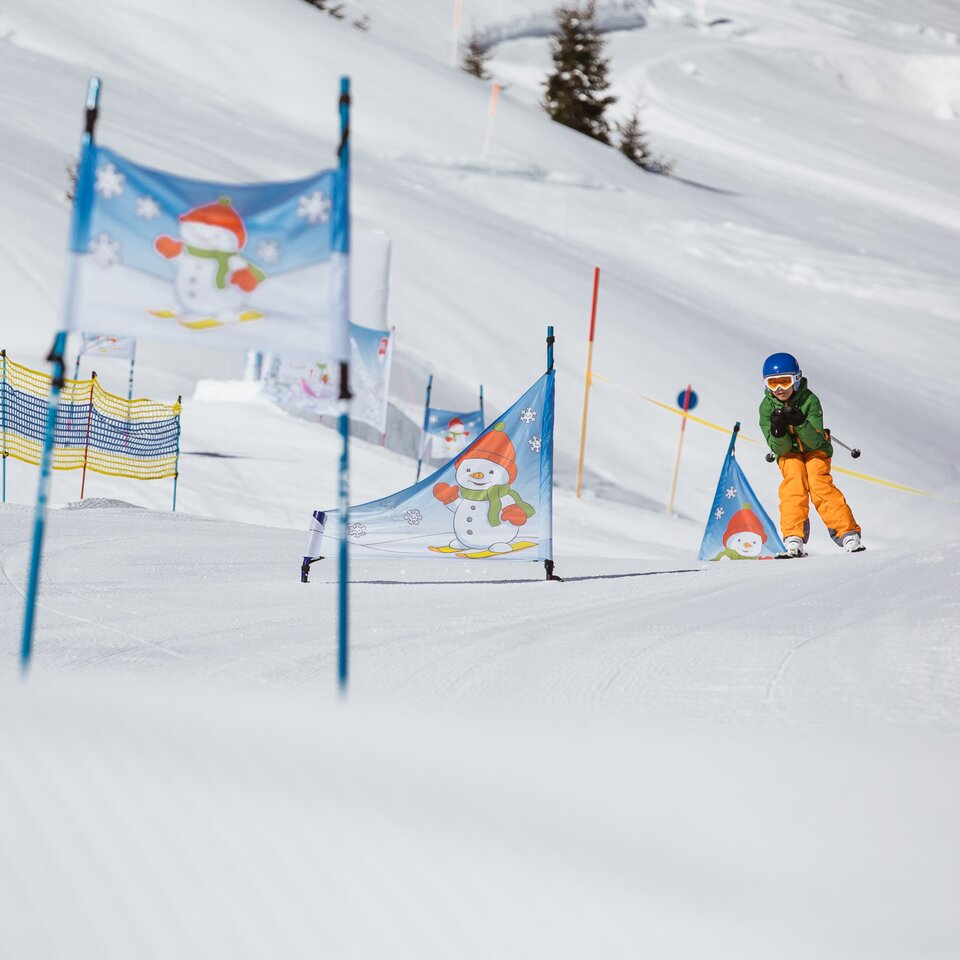 Ein Kind mit gelber Skihose fährt auf einer Piste mit Schneemann-Toren Ski.