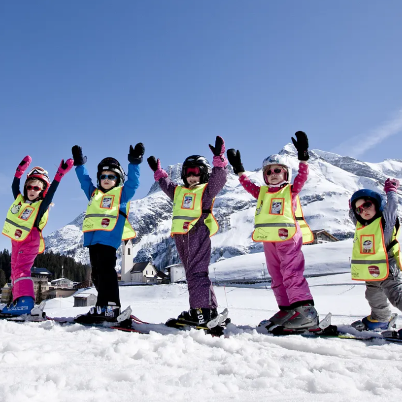 Kinder mit einer Skilehrerin in Skiausrüstung auf einer Skipiste strecken die Arme in die Luft.