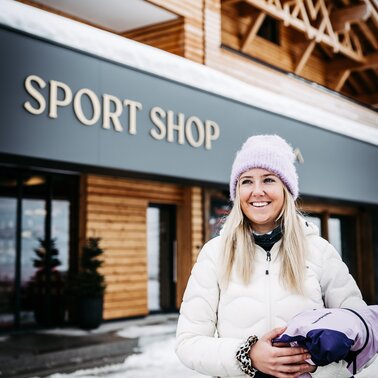 Eine lächelnde Frau in Winterkleidung steht vor einem Sportgeschäft.