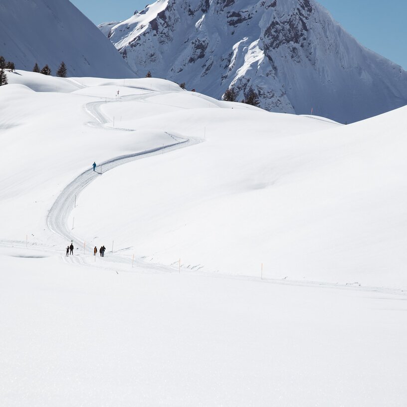 Menschen wandern auf einem verschneiten Bergweg in Richtung eines Berggipfels.