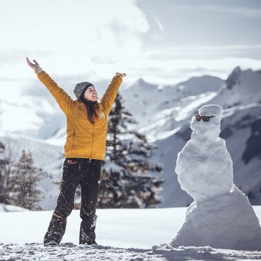 A person with arms raised in joy near a snowman in sunglasses.